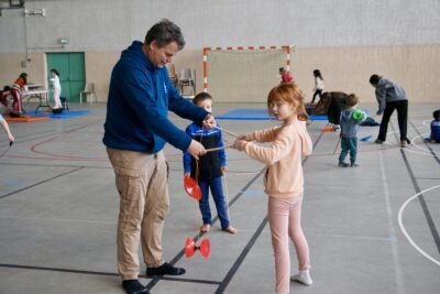 un homme montre à une jeune fille comment faire du diabolo, un autre jeune garçon regarde, à l'arrière plan on voit d'autres enfants sur d'autres activités dans le gymnase, avec les animateurs - Agrandir l'image 11 sur 16, fenêtre modale