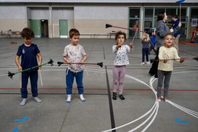 quatre enfants dans un gymnase jouent avec ce qui ressemble à un baton de majorette qu'ils tiennent en équilibre à l'horizontale à l'aide de deux autres batons - Agrandir l'image 10 sur 16, fenêtre modale