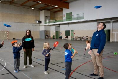 photo de plusieurs enfants dans le gymnase qui regardent impressionnés un homme qui fait tenir en équilibre un baton avec au bout d'une assiette sur son menton - Agrandir l'image 7 sur 16, fenêtre modale