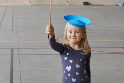 photo d'une jeune fille dans un gymnase qui fait tourner une assiette en plastique bleue en équilibre sur un piquet en bois, elle en porte une sur sa tête aussi et sourit à l'objectif - Agrandir l'image 6 sur 16, fenêtre modale