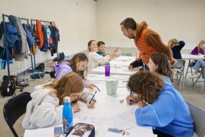 dans une salle, on voit une dizaine d'enfants répartis sur des tables, en train de dessiner sur des feuilles blanches, accompagnés par un homme avec une doudoune orange - Agrandir l'image 4 sur 10, fenêtre modale