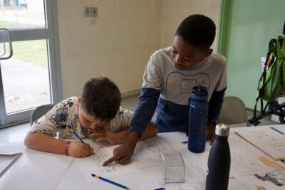 deux jeunes garçons derrière une table, un debout montre quelque chose sur la feuille d'un autre qui est assis en train de dessiner - Agrandir l'image 5 sur 10, fenêtre modale