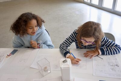 deux jeunes filles assises à une table dessinent sur une feuille blanche dans des cases - Agrandir l'image 7 sur 10, fenêtre modale