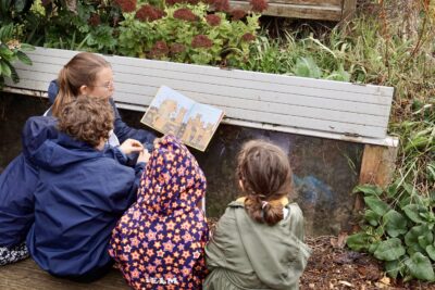 photo d'enfants qui lisent un livre pour enfant dans un espace végétalisés, assis devant une sorte de jardinière - Agrandir l'image 1 sur 14, fenêtre modale