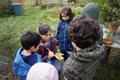 une femme montre quelque chose dans sa main à cinq enfants autour d'elle, ils sont dans ce qui pourrait être un grand jardin - Agrandir l'image 9 sur 14, fenêtre modale