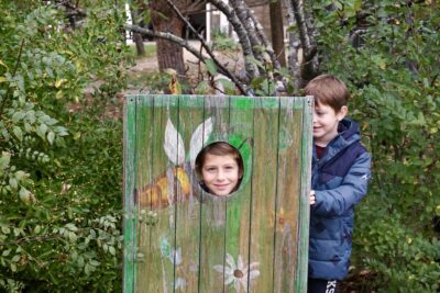 un enfant a sa tête dans un trou dans un assemblage de planches peintes, il sourit pour la photo, un enfant à côté regarde - Agrandir l'image 12 sur 14, fenêtre modale