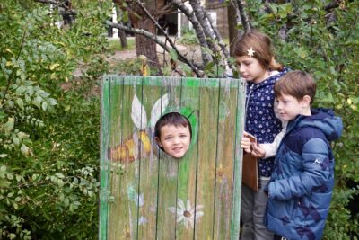 un enfant a sa tête dans un trou dans un assemblage de planches peintes, il sourit pour la photo, deux enfants à côté regardent - Agrandir l'image 13 sur 14, fenêtre modale