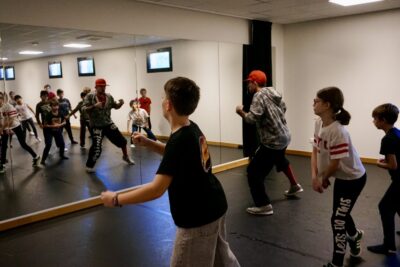 photo d'une dizaine d'enfants devant la glace dans une salle de danse, avec un homme adulte qui montre des pas - Agrandir l'image 5 sur 9, fenêtre modale