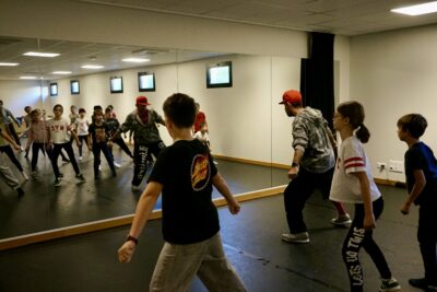 photo d'une dizaine d'enfants devant la glace dans une salle de danse, avec un homme adulte qui montre des pas - Agrandir l'image 6 sur 9, fenêtre modale