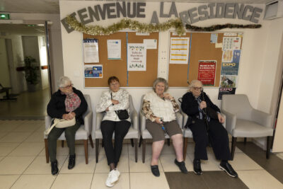 quatre femmes âgées sont assises dans des fauteuils contre un mur, elles sourient à l'objectif, au-dessus d'elles, on voit une guirlande dorée et des lettres qui forment les mots "bienvenue à la résidence" - Agrandir l'image 6 sur 26, fenêtre modale