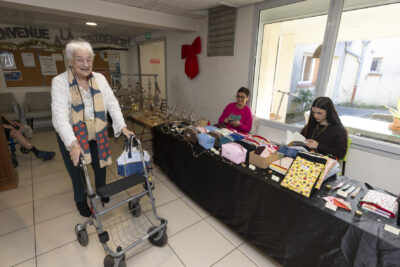 photo d'une femme âgée avec déambulateur qui fait un grand sourire à l'objectif en passant devant un stand de vente de sacoches - Agrandir l'image 21 sur 26, fenêtre modale