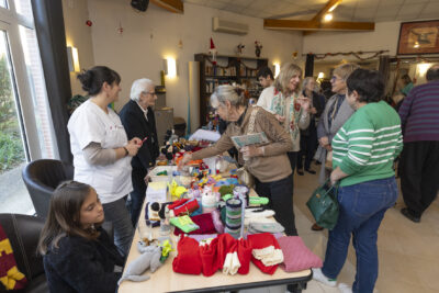 photo d'un stand vu de côté qui vend des doudous et chaussettes de Noël, une jeune fille est assise derrière, une femme avec une blouse et une femme âgée sont également derrière. Le stand est dans une salle avec une dizaine des personnes dans le champ - Agrandir l'image 4 sur 26, fenêtre modale