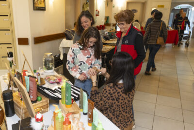 photo d'une jeune fille qui essaie un bracelet aidée par une femme accroupie, à côté d'une femme âgée et d'une autre femme souriante - Agrandir l'image 11 sur 26, fenêtre modale