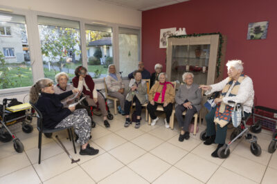 photo d'une dizaine de personnes âgées, des femmes et un homme assis en demi-cercle dans une salle, on voit le jardin de la résidence par la fenêtre derrière - Agrandir l'image 14 sur 26, fenêtre modale