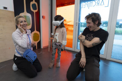 une enfant porte un casque de réalité virtuelle dans une pièce, à côté d'elle une femme et un homme souriants - Agrandir l'image 18 sur 32, fenêtre modale