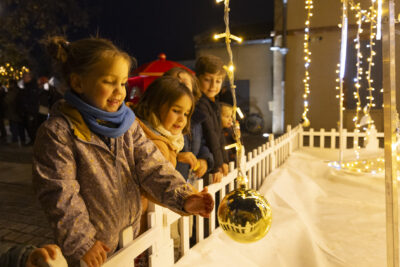 une petite fille approche la main d'une boule de Noël dorée suspendue à une guirlande lumineuse, d'autres enfants à côté la regardent - Agrandir l'image 5 sur 20, fenêtre modale