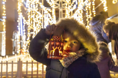 un enfant tient une petite lampe par la main, avec des trous en forme de rennes, devant son visage. à l'arrière plan en flou on voit l'illumination en forme d'arbre de la place de la Mairie - Agrandir l'image 17 sur 20, fenêtre modale