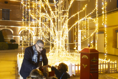 photo de nuit, le Maire et quatre enfants appuient ensemble sur un buzzer sur la place de la Mairie, derrière eux, une boîte aux lettres rouges, et une installation lumineuse en forme d'arbre d'où tombent des guirlandes dorées - Agrandir l'image 3 sur 20, fenêtre modale