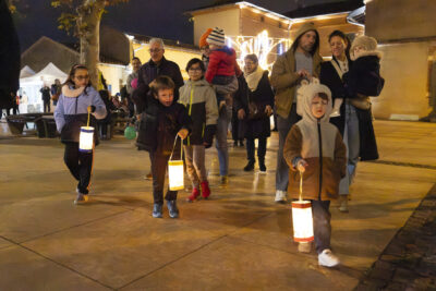 photo de la marche aux lampions, les familles marchent, les enfants au premier plan qui portent les lampions, sur la place de la mairie dans la nuit - Agrandir l'image 16 sur 20, fenêtre modale