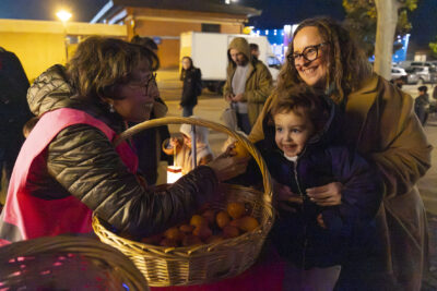 une femme et un enfant prennent une clémentine donnée par une femme qui porte un gilet rose par dessus sa doudoune - Agrandir l'image 9 sur 20, fenêtre modale