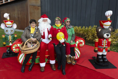 photo du Père Noël assis sur un canapé aux couleurs de Noël avec deux femmes déguisées en lutin et une autre femme qui tient un panier en osier sur ses genoux - Agrandir l'image 10 sur 12, fenêtre modale