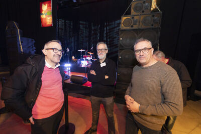 photo de trois hommes qui sourient, à côté d'une scène avec enceintes, batterie et micros positionnés - Agrandir l'image 9 sur 20, fenêtre modale